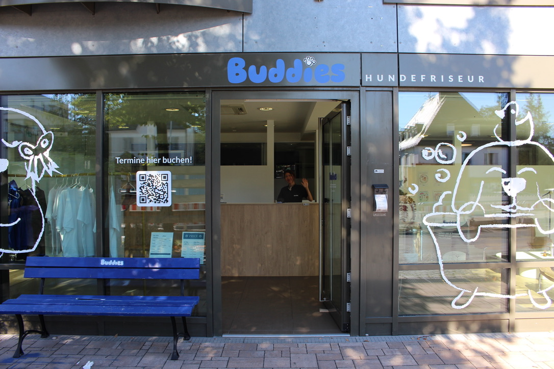 Die Buddies Storefront in Hadern: offene Tür, blaue Bank und Fensterzeichnungen